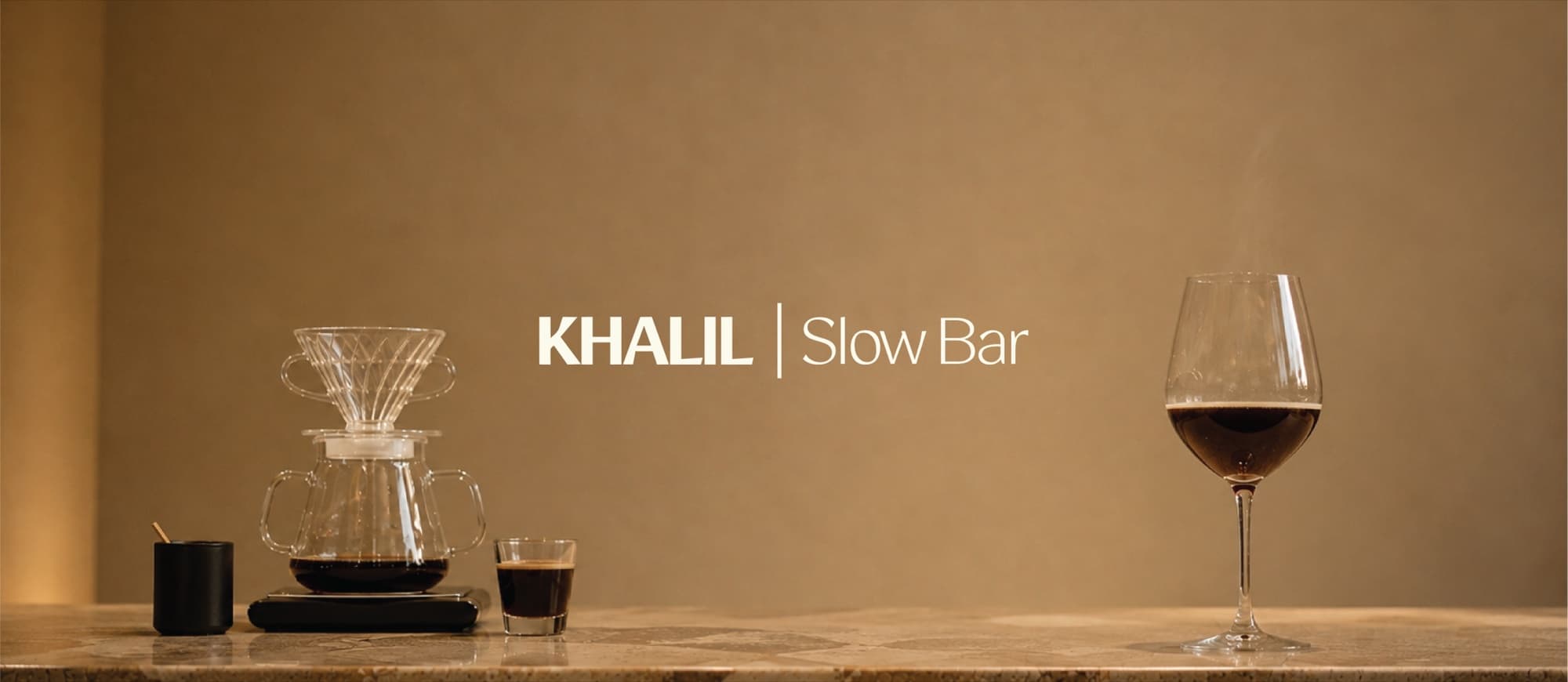 بار خليل البطيء — أدوات تقطيرٍ وكأس قهوةٍ على البار الرخامي، وفي الوسط شعار KHALIL Slow Bar